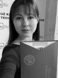 Studentė tamsiais plaukais, laiko universiteto diplomą rankose ir žiūri į kamerą.