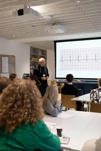 Lektorius, su akiniais ir juodu megztiniu, stovi priešais didelį ekraną, kuriame rodomas širdies elektrokardiogramos vaizdas. Auditorijoje sėdi keli moksleiviai, įdėmiai klausantys ir žiūrintys į ekraną. Aplinka primena modernią mokymo klasę ar laboratoriją, su minimalistiniu dizainu ir šiuolaikinėmis technologijomis.