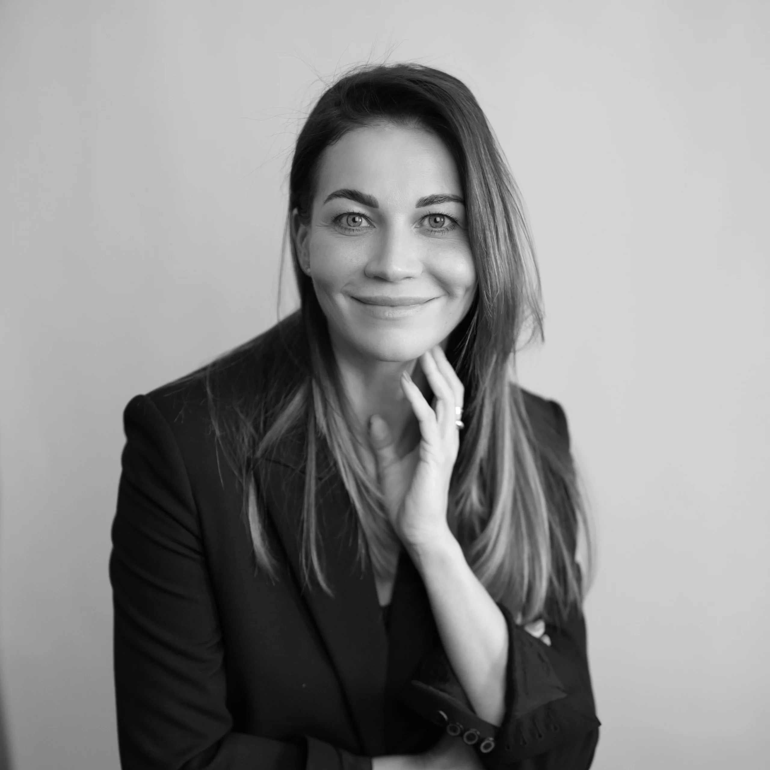 Juodai balta portretinė nuotrauka, vaizduojanti besišypsančią moterį su ilgais plaukais, vilkinčią juodą švarką. Ji žvelgia tiesiai į kamerą, švelniai prilaikydama veidą ranka.
