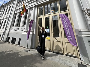 Jauna, šviesių plaukų moteris stovi prie Kauno technologijos universiteto, Socialinių, humanitarinių mokslų ir menų fakulteto pastato, šalia įėjimo durų. Greta matomos violetinės vėliavos. Nuotraukoje taip pat matomas giedras dangus ir klasikinė architektūra.