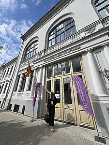 Jauna, šviesių plaukų moteris stovi prie Kauno technologijos universiteto, Socialinių, humanitarinių mokslų ir menų fakulteto pastato, šalia įėjimo durų. Greta matomos violetinės vėliavos. Nuotraukoje taip pat matomas giedras dangus ir klasikinė architektūra.