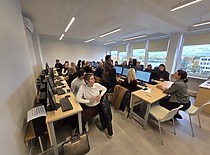 Auditorijoje matosi daug žmonių, sėdinčių ir klausiančiųsi mokymų; priekyje stovi lektorius, rodantis skaidres ar pristatymą.