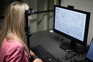 Moteris rožiniu švarku sėdi prie kompiuterio ir atlieka tyrimą žvilgsnio sekimo laboratorijoje. Prie jos galvos pritvirtinta žvilgsnio sekimo įranga, o ekrane rodomas tekstas, kurį ji skaito.