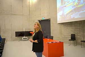 Asmuo stovi modernioje konferencijų salėje prie ryškiai oranžinio stalo, ant kurio sudėti keli bukletai ir vandens butelis. Fone matoma didelė projekcijos ekranas su spalvinga iliustracija, pilkos betoninės sienos, televizoriaus ekranas ir kelios tuščios kėdės.