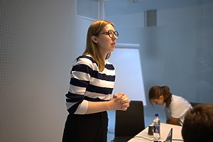 Moteris stovi prie stalo moderniame biuro kambaryje, vilkėdamas juostuotą megztinį. Ant stalo yra nešiojamasis kompiuteris, vandens butelis ir dokumentai. Fone matomas tuščias popieriaus lapas ant stovinčio stovo (flipchart) ir stiklinė pertvara.