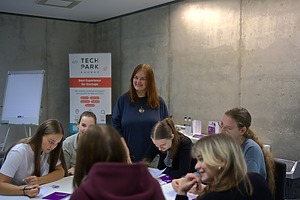 Grupė žmonių sėdi prie stalo moderniame kambaryje, ant stalo yra dokumentai ir rašymo priemonės. Fone matomas stovas su popieriaus lapu ir reklaminis stendas su užrašu „Tech Park – Best Experience for Startups“. Kambario sienos pilkos, iš betono.