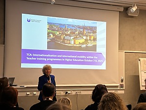 Moteris pristato renginį auditorijoje, projekcijos ekrane matomas užrašas „TCA: Internationalisation and international mobility within the Teacher training programmes in Higher Education October 7–9, 2025“, su Švedijos miestovaizdžio nuotrauka.