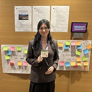 Moteris stovi prie sienos su spalvingais lipdukais ir plakatais, rankose laikydama vardinę kortelę. Ji šypsosi, vilki tamsų švarką ir stovi konferencijos aplinkoje, kur matyti informaciniai lapai ir ekrane rodoma „Swedish Council for Higher Education“ iškaba.