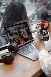 Asmuo sėdi prie stalo su atverstu nešiojamuoju kompiuteriu „MacBook Air“, kuriame matomos nuotraukos. Šalia guli ausinės, o rankose laikoma fotoaparatą, jungiant jį prie kompiuterio vaizdų peržiūrai ar redagavimui. Fone matosi žmonės už stiklo.