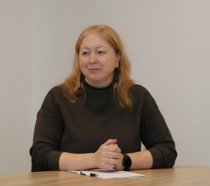 Moteris sėdi prie medinio stalo, sukryžiavusi rankas. Ji dėvi tamsų megztinį, turi šviesius banguotus plaukus ir žiūri šiek tiek į šoną. Ant stalo prieš ją padėtas lapas su rašikliu. Fone – šviesi, neutrali siena.