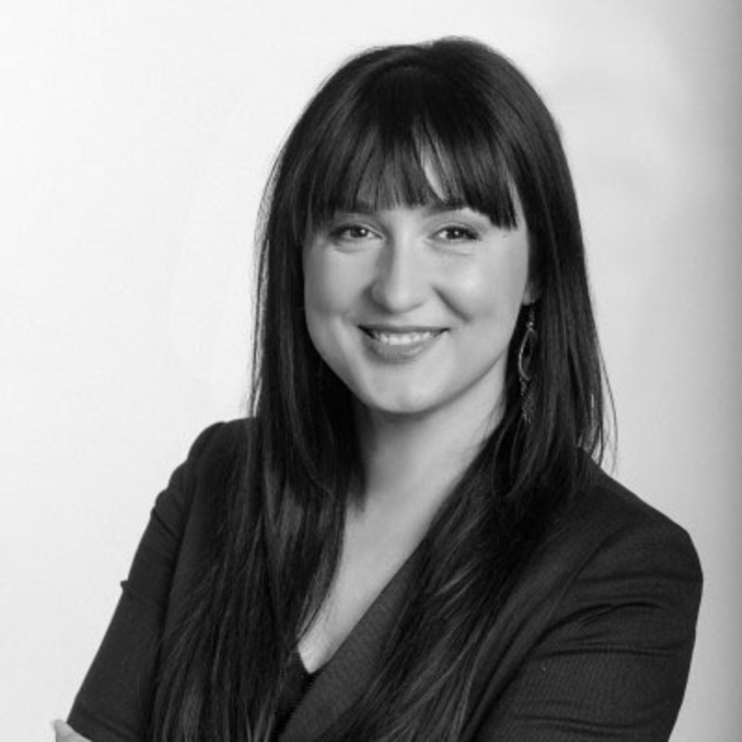 Juodai balta portretinė nuotrauka, kurioje moteris su ilgais tamsiais plaukais ir kirpčiukais šypsosi, apsirengusi švarku ir sukryžiavusi rankas.