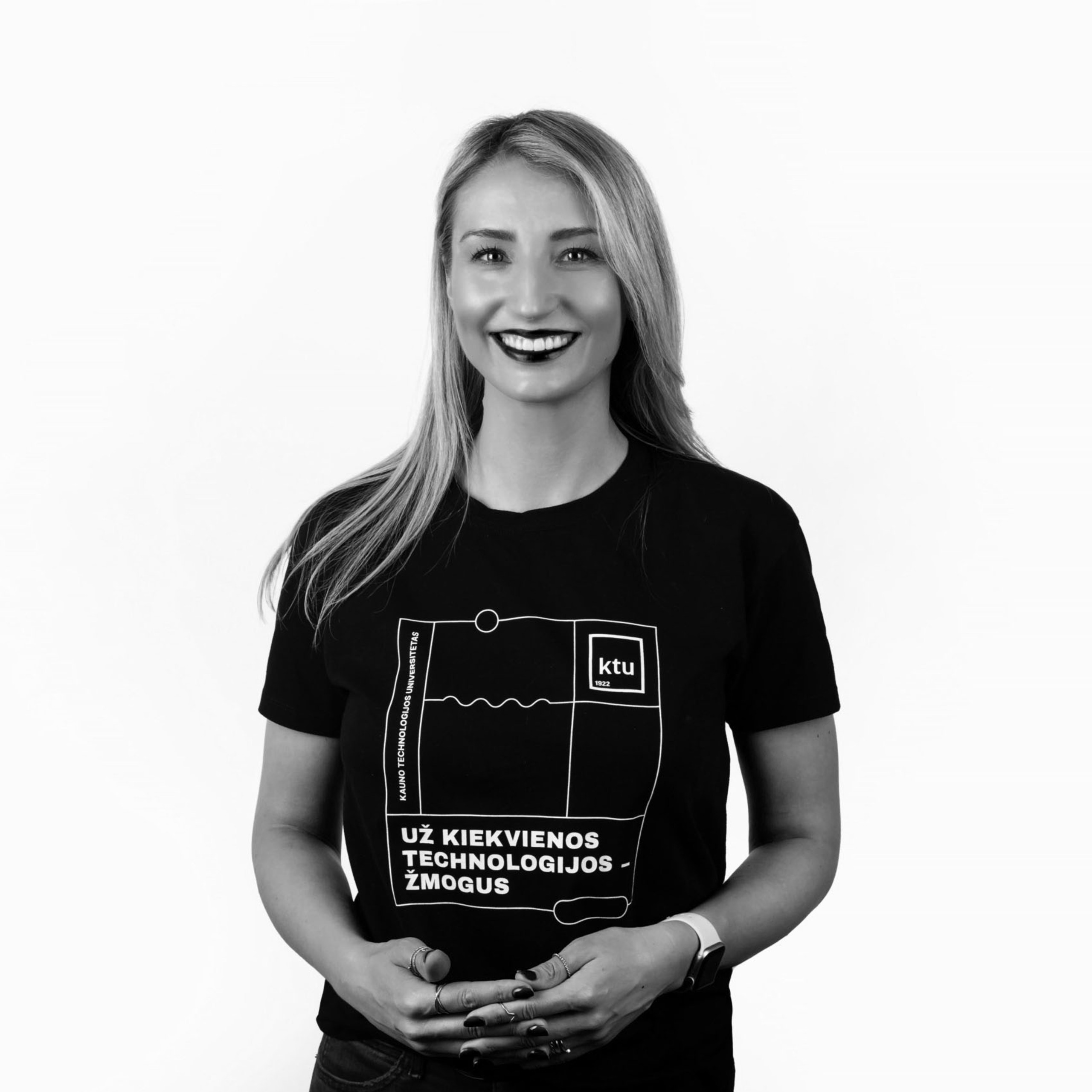 Juodai balta nuotrauka, kurioje jauna moteris šypsosi ir pozuoja prieš šviesų foną, vilkėdama juodą KTU marškinėlių su užrašu „Už kiekvienos technologijos – žmogus“.