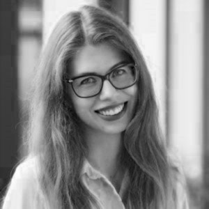 Juodai baltas jaunos moters portretas: ji šypsosi, vilki šviesius drabužius ir dėvi akinius, o ilgi plaukai laisvai krinta ant pečių.