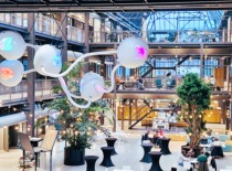 Šiuolaikinė bendradarbiavimo erdvė su industrinio stiliaus architektūra, stiklinėmis sienomis ir menine šviesos instaliacija, kurioje žmonės dirba ir diskutuoja kūrybiškoje, inovatyvioje aplinkoje.“