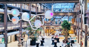 Šiuolaikinė bendradarbiavimo erdvė su industrinio stiliaus architektūra, stiklinėmis sienomis ir menine šviesos instaliacija, kurioje žmonės dirba ir diskutuoja kūrybiškoje, inovatyvioje aplinkoje.“