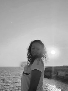 Mergina garbanotais plaukais stovi prie vandens telkinio saulėlydžio metu, vilkėdama &scaron;viesius mar&scaron;kinėlius. Fone matyti ramus jūros ar vandenyno pavir&scaron;ius ir tolumoje esanti žemė ar uolos.