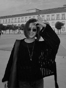 oteris su akiniais nuo saulės ir megztu juodu megztiniu &scaron;ypsosi ir taiso plaukus vėjuotą dieną miesto aik&scaron;tėje su istoriniu pastatu fone.