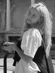 Mergina ilgais plaukais stovi lauke prie&scaron;ais pastatą su klasikinės architektūros elementais &ndash; kolonomis ir faktūrine siena. Ji vilki &scaron;viesius mar&scaron;kinėlius, rankose laiko daiktą, galimai telefoną, o per petį persimetusi tamsi rankinė.