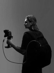 Vaikinas su kuprine laiko fotoaparatą ant stabilizatoriaus ir žvelgia per petį. Nuotrauka daryta tamsiame fone, pabrėžiant filmavimo procesą.