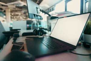 Nešiojamas kompiuteris su tuščiu ekranu ant biuro stalo šalia išorinio monitoriaus, ausinių, telefono ir augalo modernioje atviro plano darbo erdvėje.