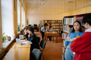 Bibliotekoje studentai mokosi: vieni dirba prie kompiuterių, kiti skaito ar diskutuoja prie stalų ir knygų lentynų.