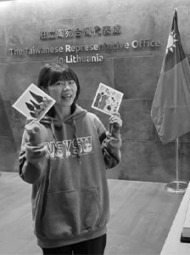 Mergina su džemperiu stovi prie sienos su užra&scaron;u &bdquo;The Taiwanese Representative Office in Lithuania&ldquo;, rankose laiko du atvirukus. &Scaron;alia matoma Taivano vėliava.